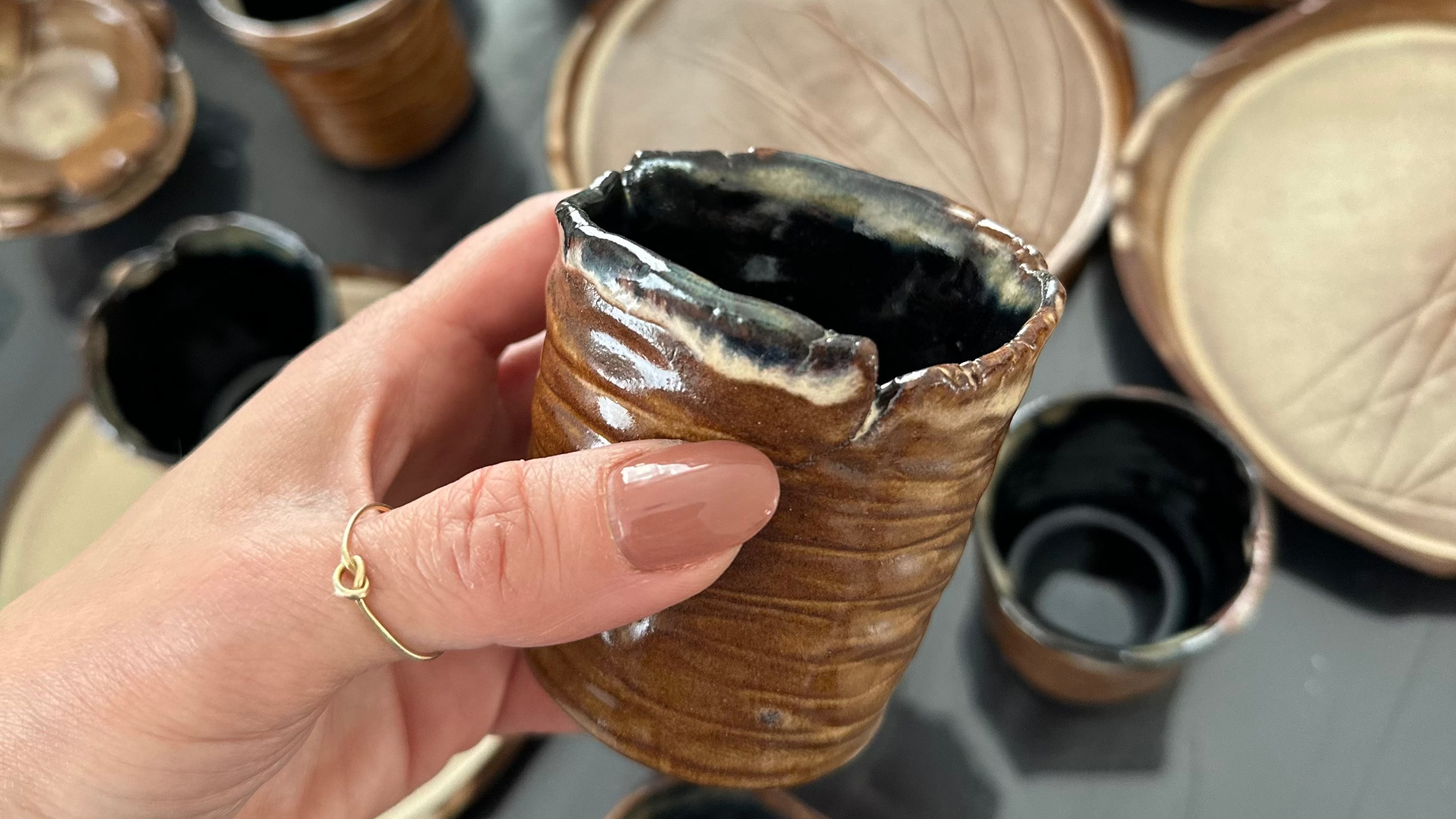 Hand holding a small ceramic cup with a textured surface, surrounded by more ceramic cups on a reflective surface.